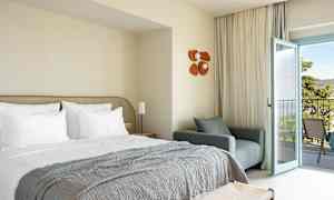 Hotelværelse med redt seng, lænestol og åben altandør. (AI-genereret tekst)