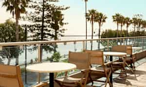 Træstole og borde på en altan med udsigt over palmer og hav. (AI-genereret tekst)