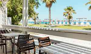 En terrasse med borde og stole, med udsigt over en grøn græsplæne, palmer og stranden i baggrunden. (AI-genereret tekst)