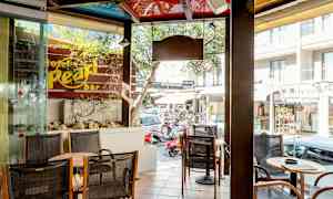 Et caféområde med siddepladser og borde ved siden af en butiksrude. Udenfor ses en gade med butikker og biler. (AI-genereret tekst)