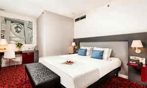 Et hotelværelse med en stor seng, to blå puder, et skrivebord med stol, rødt tæppe og moderne lamper. (AI-genereret tekst)