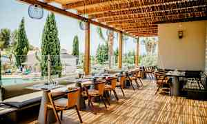 Restaurant udendørs terrasse med borde og stole, med udsigt over en pool og træer. (AI-genereret tekst)