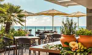 En udendørs terrasse med borde og stole under parasoller, med havet i baggrunden og grønne planter samt eksotiske frugter på et bord i forgrunden. (AI-genereret tekst)