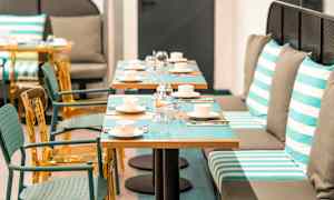 Billede af en restaurant med borde dækket med kopper, glas og bestik. Stole og bænkepladser med stribede puder ses. (AI-genereret tekst)