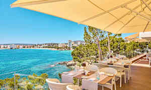 Udendørs servering med udsigt over en klarblå havbugt og bygninger i det fjerne. (AI-genereret tekst)