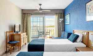 Billede af et hotelværelse med to senge, en blå væg og en glasdør mod en altan. (AI-genereret tekst)