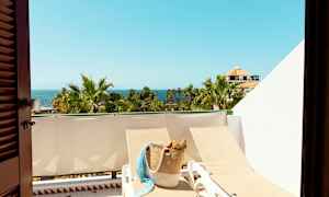 Udsigt over en solrig altan med liggestole, en kurvtaske, palmer og havet i baggrunden. (AI-genereret tekst)