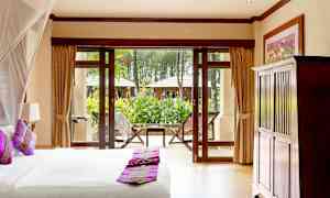 Et soveværelse med en seng og puder i lilla farver, udsigt til en have gennem store vinduer med døre. (AI-genereret tekst)