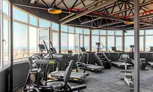 Lyst fitnesscenter med træningsmaskiner og udsigt gennem store vinduer. (AI-genereret tekst)