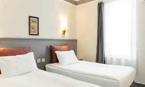 To senge med hvidt sengetøj og røde puder står i et rum med lyse vægge og en væglampe. Et vindue med hvide gardiner er til højre. (AI-genereret tekst)