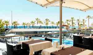 Udsigt fra en udendørs servering mod et poolområde med parasoller og palmer samt havet i baggrunden. (AI-genereret tekst)