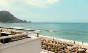 Et billede af en strand med liggestole, parasoller og mennesker, der svømmer i havet. I baggrunden ses en klippe. (AI-genereret tekst)