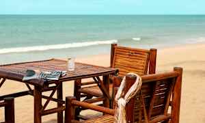 Stole og bord af træ står på en strand med havet i baggrunden. (AI-genereret tekst)