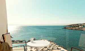 En balkon med udsigt over havet, parasoller ved en pool og en kop på et bord. (AI-genereret tekst)