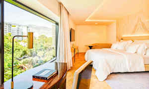 Et hotelværelse med en stor seng og et skrivebord nær et stort vindue, der viser udsigten over byen. (AI-genereret tekst)