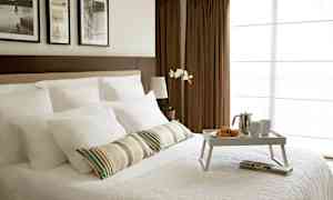 Et værelse med en dobbeltseng redt med hvidt sengetøj og en morgenmadsbakke med croissanter, en kaffekande og kopper. I baggrunden ses brune gardiner og billeder på væggen. (AI-genereret tekst)