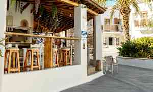 En udendørs bar med træstole og tag af terracottafliser. En palme og bygninger ses i baggrunden. (AI-genereret tekst)