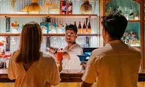 To personer sidder på en bænk ved en bardisk, der er dekoreret med flasker og farverige detaljer. (AI-genereret tekst)