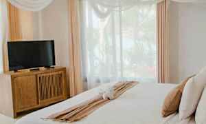 Et lyst soveværelse med en dobbeltseng med hvidt sengetøj, et TV på en trækommode, store vinduer og gardiner samt dekorative loftstekstiler. (AI-genereret tekst)