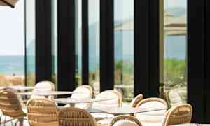 En udendørs terrasse med borde og stole ved siden af en bygning med store glasvinduer. (AI-genereret tekst)