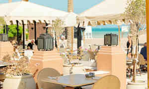 Udendørs servering med borde og stole under parasoller tæt på stranden. (AI-genereret tekst)