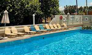 En udendørs swimmingpool med liggestole og parasoller ved siden af. Træer og en klar blå himmel i baggrunden. (AI-genereret tekst)