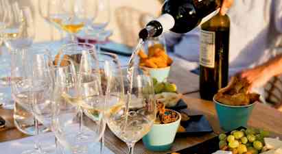 Et bord med glas fyldt med hvidvin, der serveres, omgivet af forskellige snacks som ost, brød og frugt. (AI-genereret tekst)