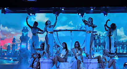 Dansere poserer på en oplyst scene med dekoration af palads-lignende bygninger mod en skyet himmel. (AI-genereret tekst)