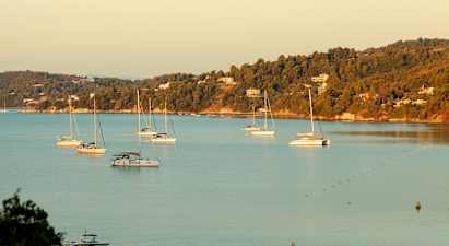 Flere sejlbåde i en bugt med skovklædte bakker og huse i baggrunden. (AI-genereret tekst)