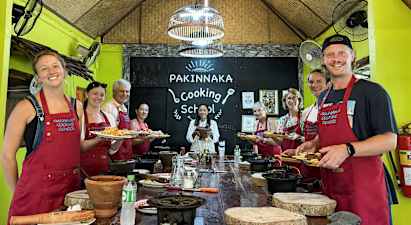 En gruppe personer iført forklæder står foran et skilt med teksten 'Pakinnaka Cooking School' i et køkkenstudio. (AI-genereret tekst)