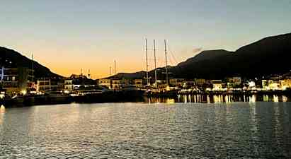 En havn i skumringen med både fortøjet ved en kaj og oplyste bygninger i baggrunden. Bjerge ses i baggrunden. (AI-genereret tekst)
