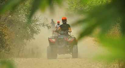Personer, der kører firhjulede køretøjer på en støvet grusvej omgivet af vegetation. (AI-genereret tekst)