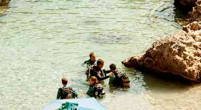 Gruppe af personer i våddragter og dykkerudstyr står i klart vand ved klipper. (AI-genereret tekst)