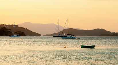 Billede af en stille bugt ved solnedgang med sejlbåde og en grøn båd i forgrunden. (AI-genereret tekst)