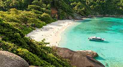 En sandstrand med mennesker, omgivet af frodige træer og klart turkis vand. En båd ligger tæt på stranden. (AI-genereret tekst)