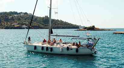 En sejlbåd med flere personer om bord sejler tæt på en kystlinje med grønne bakker og nogle huse. (AI-genereret tekst)