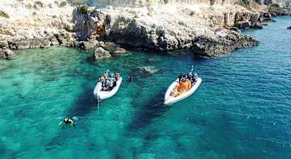 To både ligger for anker i klart turkisblåt vand nær en klippefyldt kyst. Dykkere er i vandet. (AI-genereret tekst)