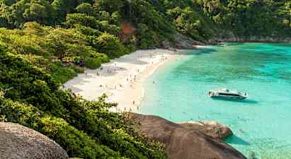 En sandstrand med mennesker, omgivet af frodige træer og klart turkis vand. En båd ligger tæt på stranden. (AI-genereret tekst)