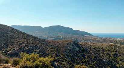 En udsigt over et bjergrigt landskab med varieret vegetation og havet i baggrunden under en klar blå himmel. (AI-genereret tekst)