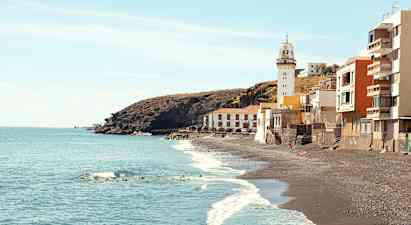 Billede af en kyst med sorte sandstrande, et fyrtårn og bygninger ved vandkanten. (AI-genereret tekst)