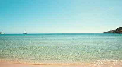 Strand med klart vand og to sejlbåde på havet. (AI-genereret tekst)