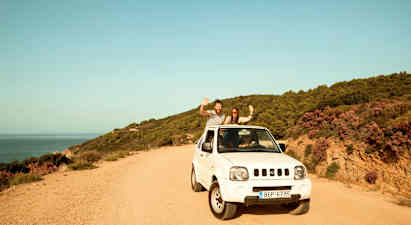 En hvid bil parkeret på en grusvej ved siden af en kystlinje med skovklædte bakker. (AI-genereret tekst)