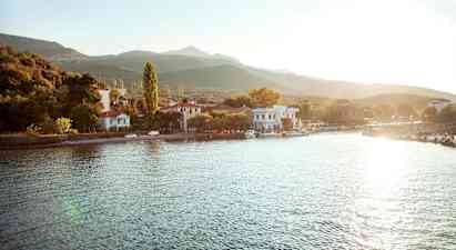 En lille landsby ligger ved en vandkant med huse og træer omgivet af bjerge. Solen skinner over området. (AI-genereret tekst)