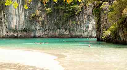 Billedet viser en strand med klart turkis vand omgivet af høje klippeformationer dækket af vegetation. Flere personer svømmer i vandet under en klar himmel. (AI-genereret tekst)