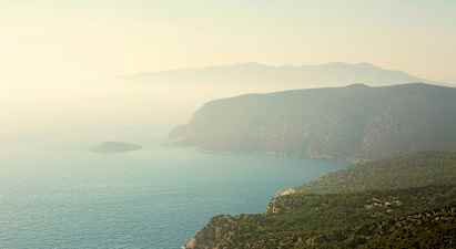 En kystlinje med stejle klipper og grønne skove, der skråner ned mod havet. En dis dækker landskabet. (AI-genereret tekst)