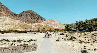 Et tørt landskab med personer, der går på en sti, der fører mod nogle små bygninger. Bjerget kan ses i baggrunden under en klarblå himmel. (AI-genereret tekst)