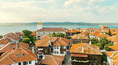 Den gamle bys tage med huse med røde teglstenstage og en kirke i baggrunden nær havet ved kysten. (AI-genereret tekst)