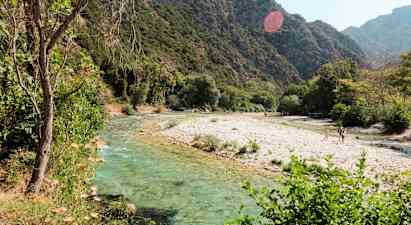 En flod med klart vand, der løber gennem et frodigt landskab med bjerge i baggrunden. (AI-genereret tekst)