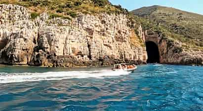 En motorbåd med mennesker sejler på blåt vand tæt på en klippe med en stor grotteåbning. (AI-genereret tekst)