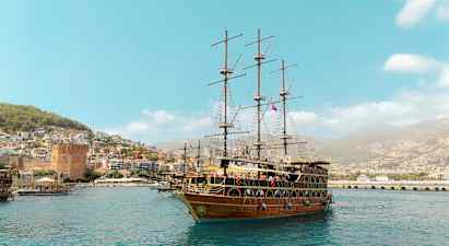 Et stort sejlskib ligger i havnen med en by og grønne bakker i baggrunden. (AI-genereret tekst)